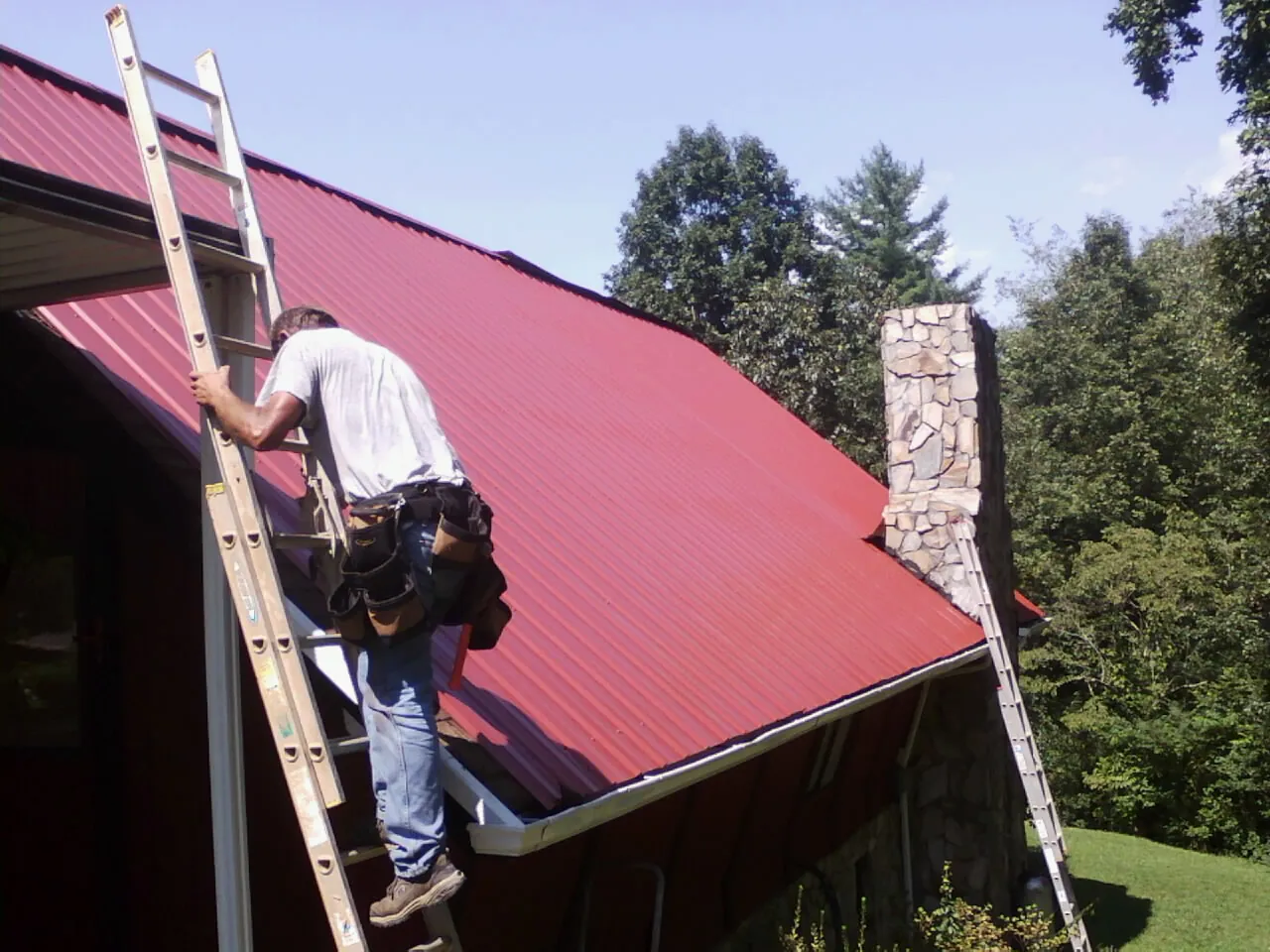 Roofer on a ladder beside a red metal roof and stone chimney — Soffit & Fascia Repair by Roofing Brothers in Fort Campbell